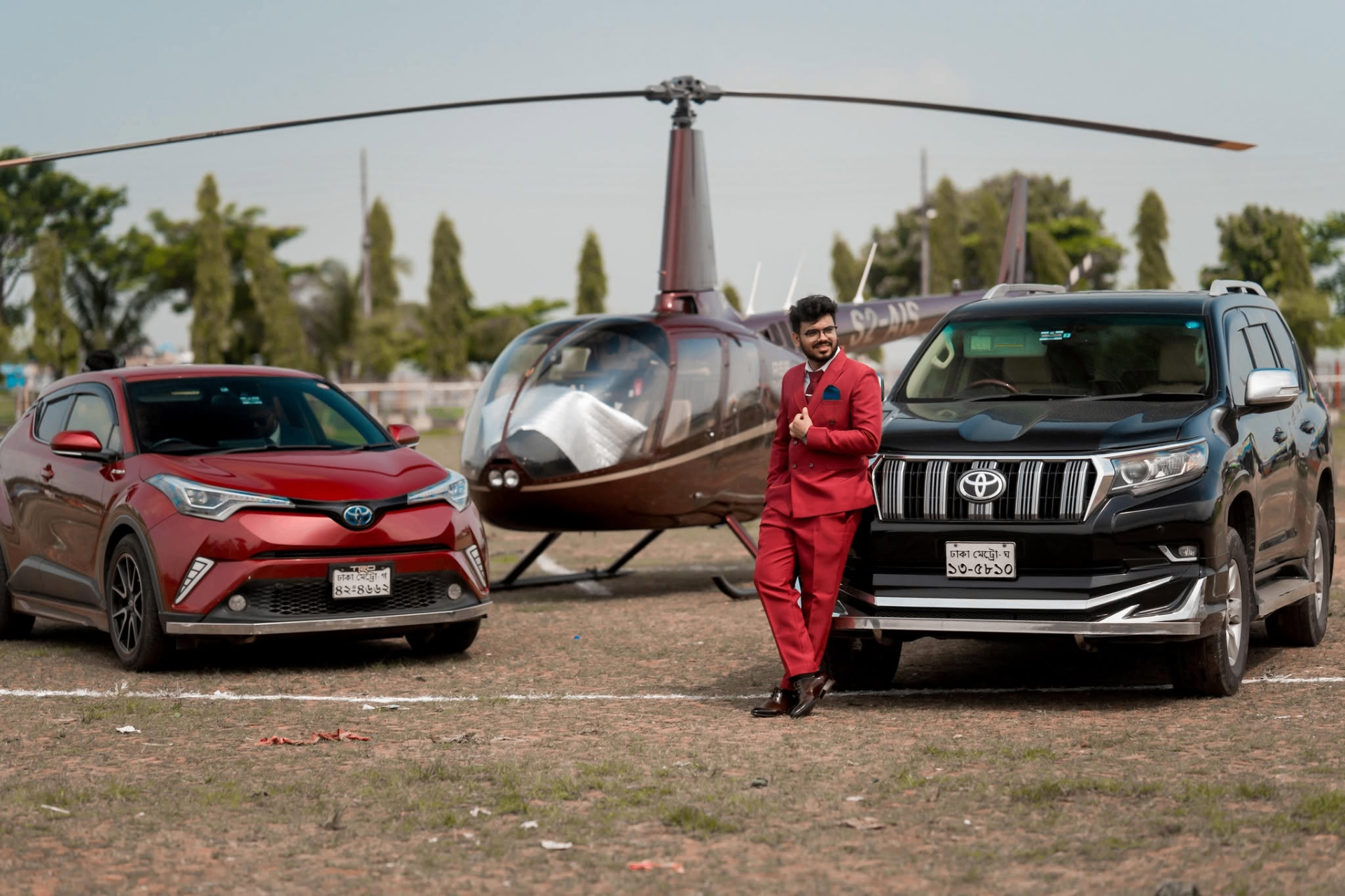 Nerob Pikash standing confidently in front of a helicopter and luxury vehicles